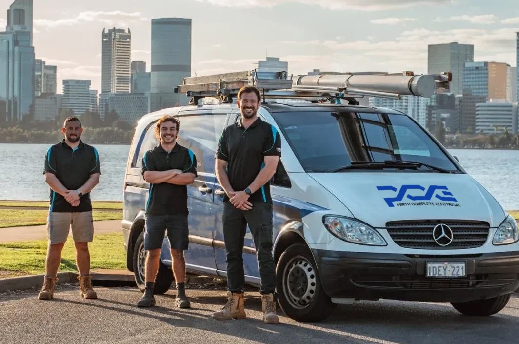 Electricians stand proudly beside their van with a city skyline in the background.